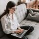A woman with glasses sitting cross legged on a gray couch working on laptop